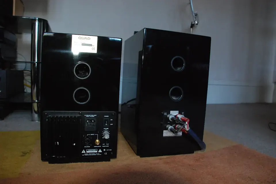 A pair of small glossy black loudspeakers and a compact subwoofer on a carpet in an empty room