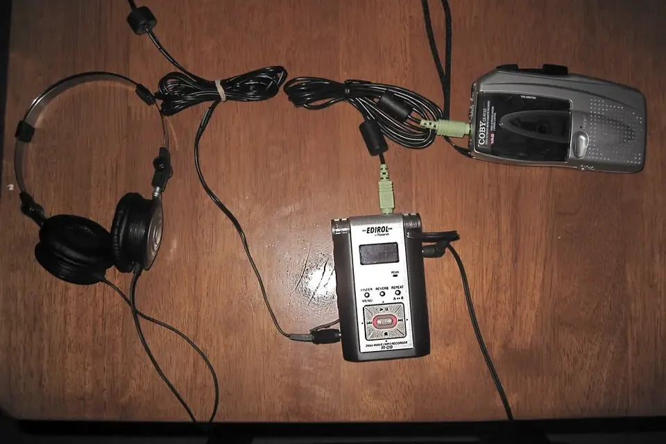 A portable cassette player connected by a cable to a small digital recorder, with headphones, on a wooden table