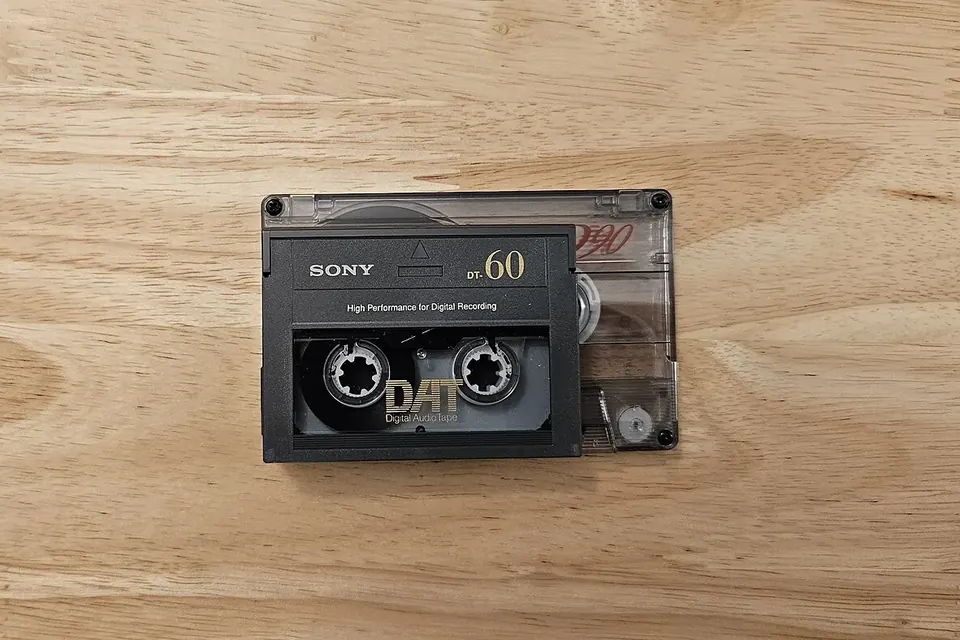 A small DAT cassette lying on top of a larger Compact Cassette on a wooden table