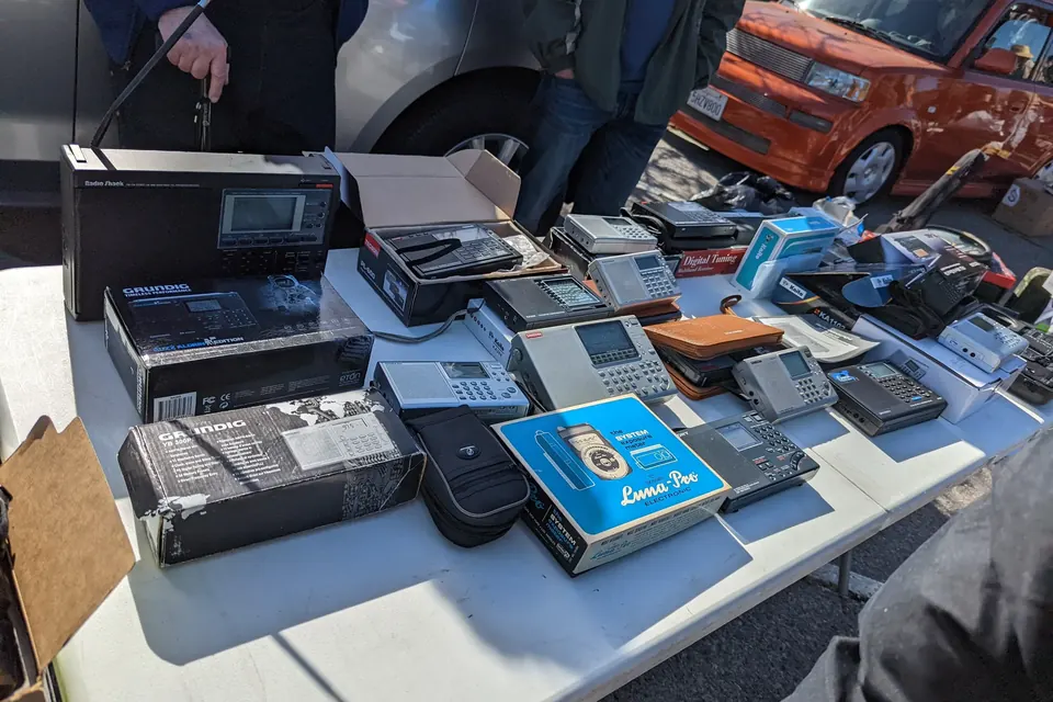 A folding table at an outdoor flea market covered with boxed and unboxed vintage portable electronics