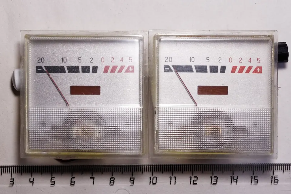 A pair of loose analogue VU meters with white scales and red overload zones, lying on a ruler