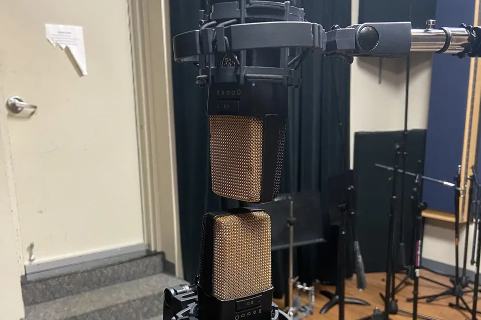 Two large studio microphones mounted one above the other on a single stand, set up as a crossed pair in a rehearsal room
