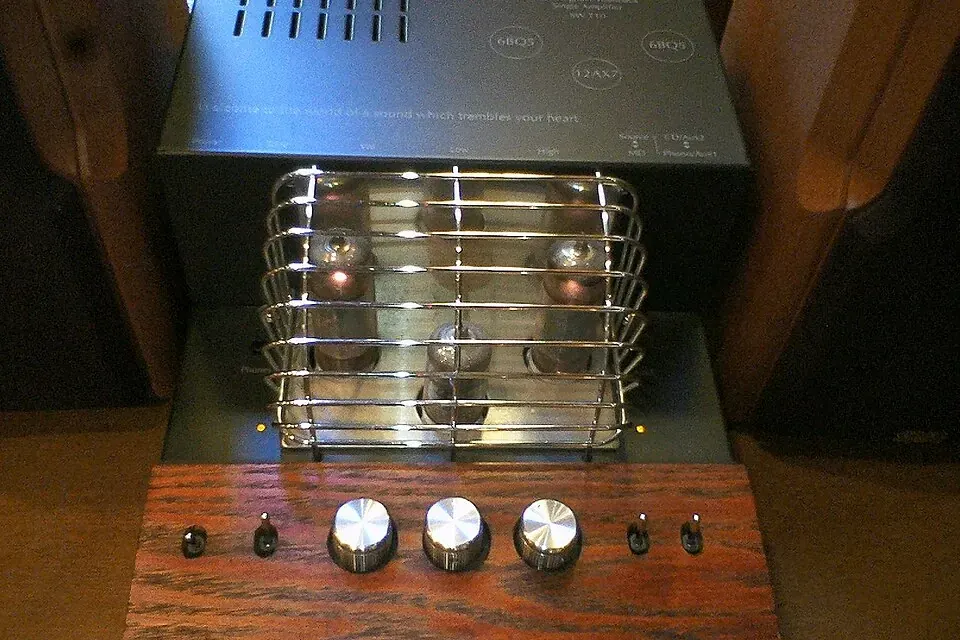 A small tube amplifier with its glass tubes inside a chrome wire cage, above a wooden front panel with three knobs