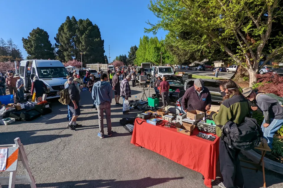 Crowds walking between tables of second-hand electronics at an outdoor flea market in a car park